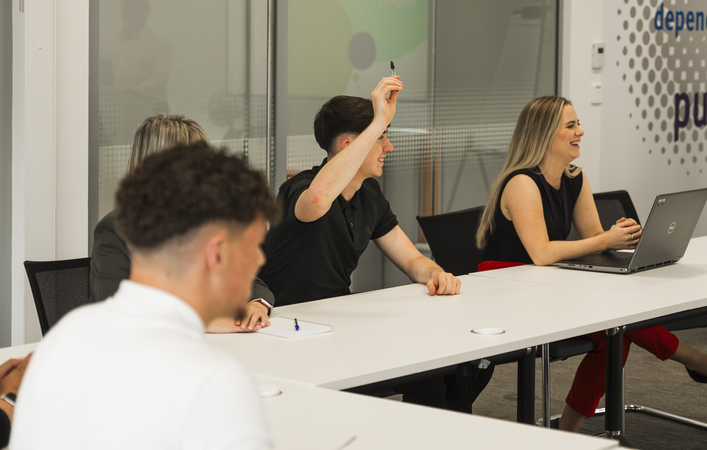 Two women, one of which obscured, and two young men, one with his hand raised, seated around a conference table.