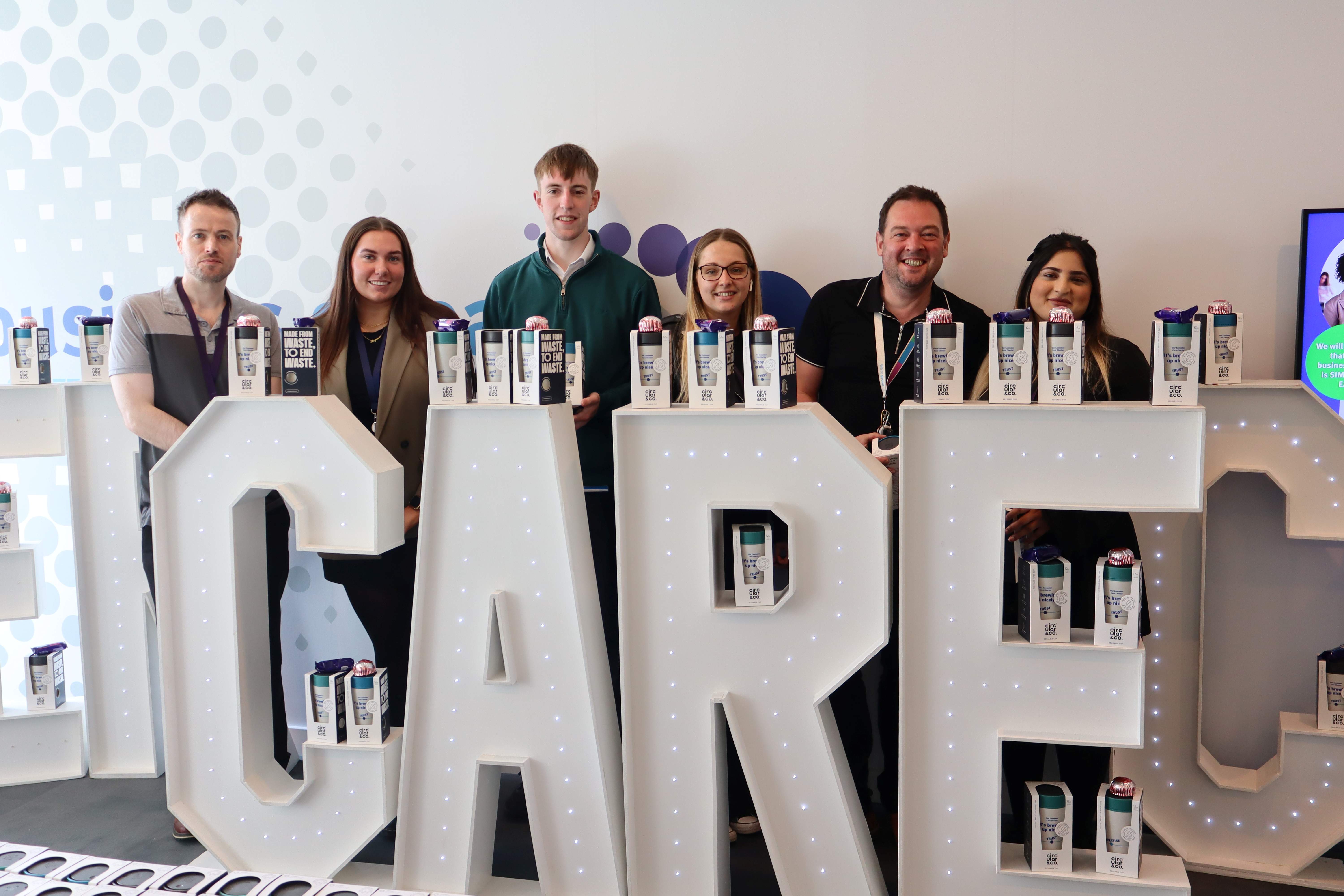 Six colleagues standing behind large floor-standing letters spelling out the world CARE