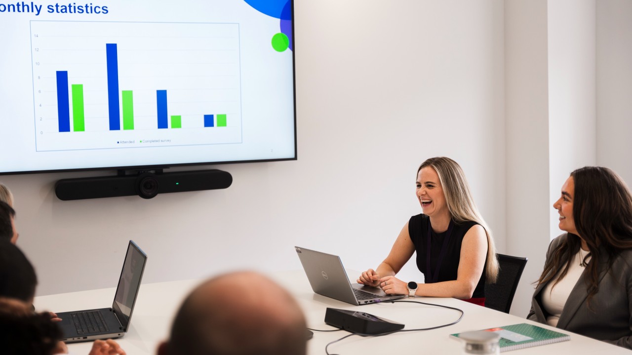 People in a meeting room with laptops, facing a screen showing a ‘Monthly statistics’ bar chart