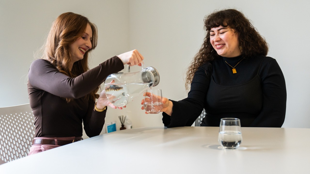 Two people at a table pouring drinking water from a glass jug into a tumbler