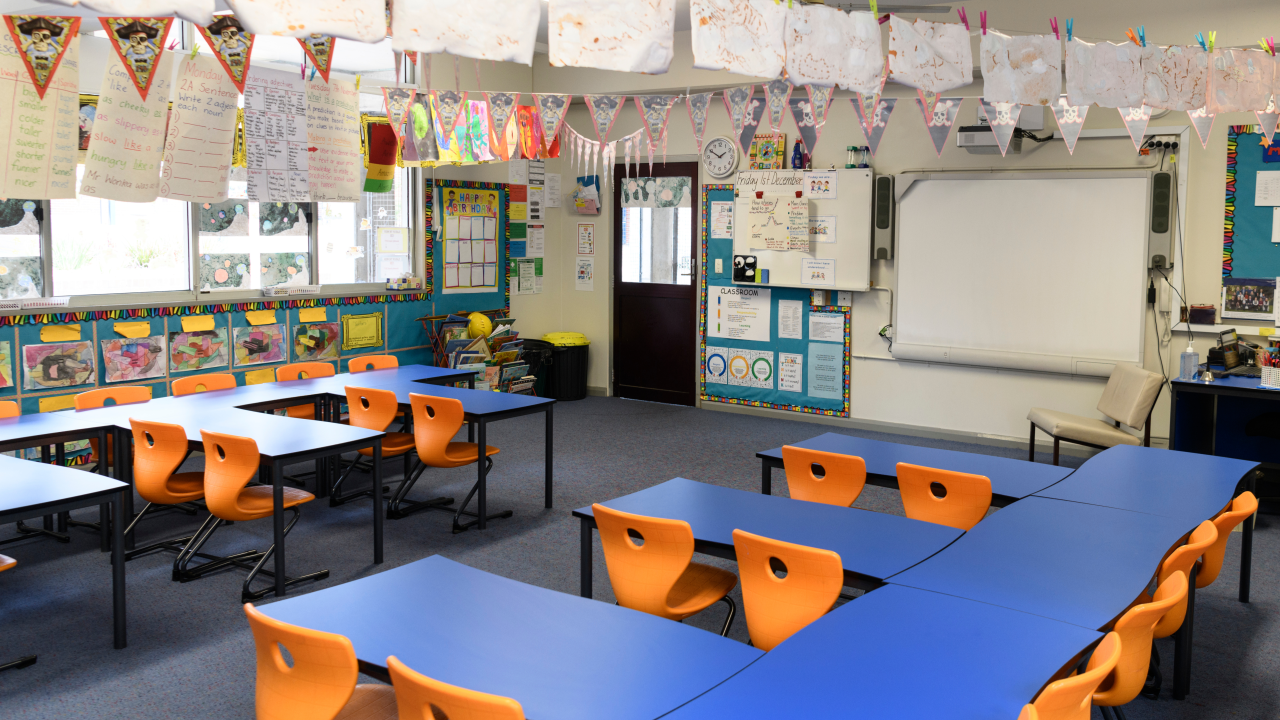 Empty school classroom with blue tables, orange chairs, colourful student artwork and learning displays decorating the walls and ceiling.