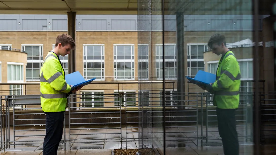 Person in a high-visibility jacket reviewing documents beside a glass building, with their reflection visible in the window.