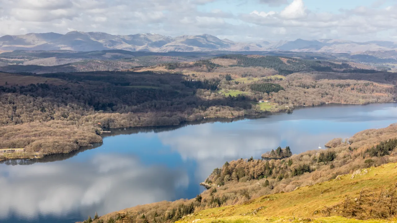 Lake surrounded by wooded hills with distant mountains under a partly cloudy sky