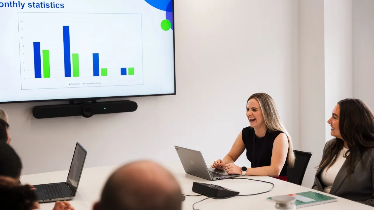People in a meeting room with laptops, facing a screen showing a ‘Monthly statistics’ bar chart