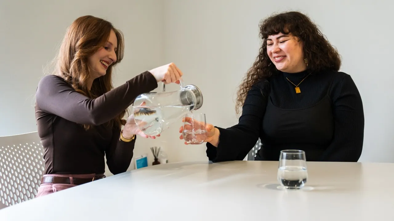 Two people at a table pouring drinking water from a glass jug into a tumbler