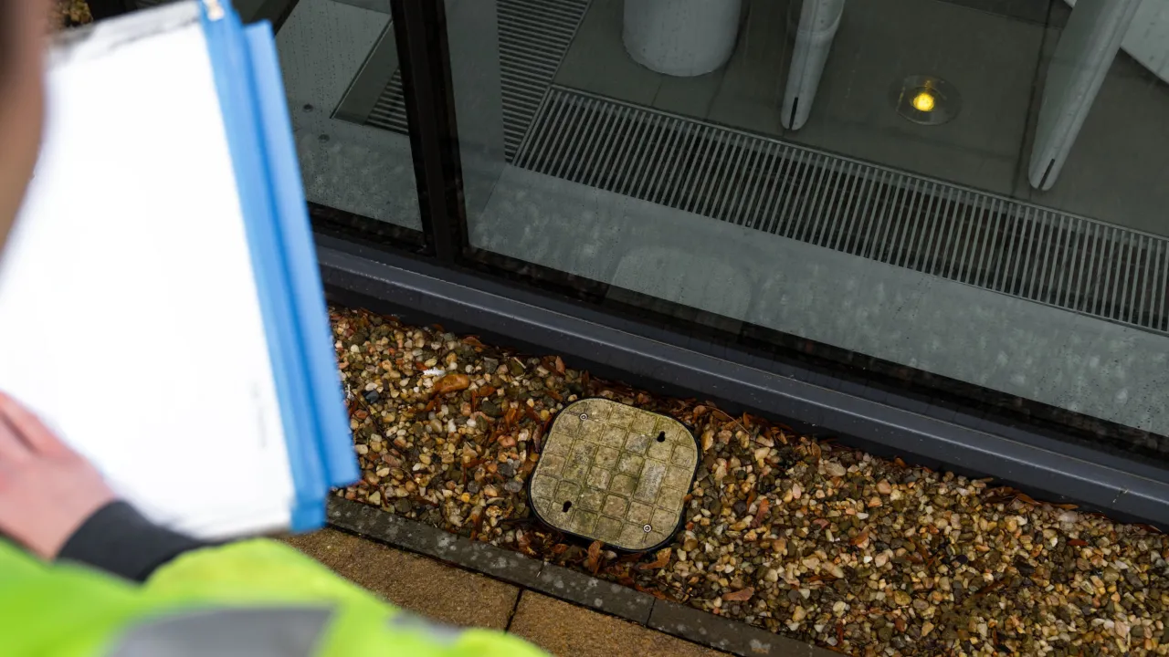 Person holding a clipboard stands by a building window, looking at a ground utility access cover set in gravel