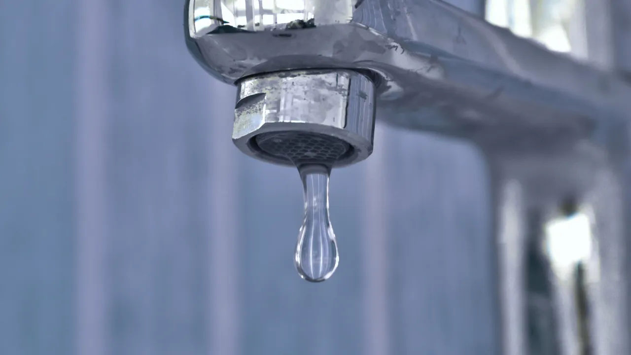 Close-up of a chrome tap with a single droplet of water hanging from the spout, set against a softly blurred blue background.