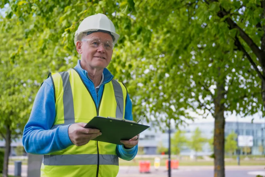 Man in white hard hat and hi-vis holding clipboard with green trees and office buildings in background