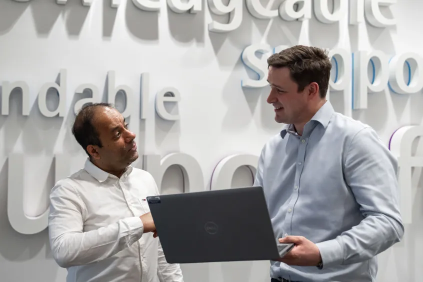 Two men standing with a laptop and talking in front of a word wall with the words knowledgeable and supportive visible