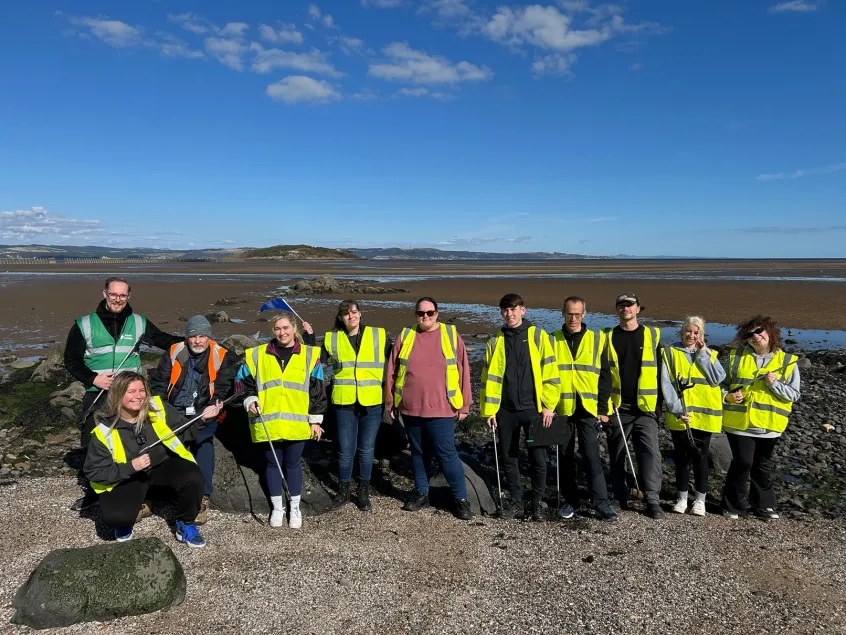 A group of 11 smiling people on a beach, wearing hi-vis vests and holding litter pickers