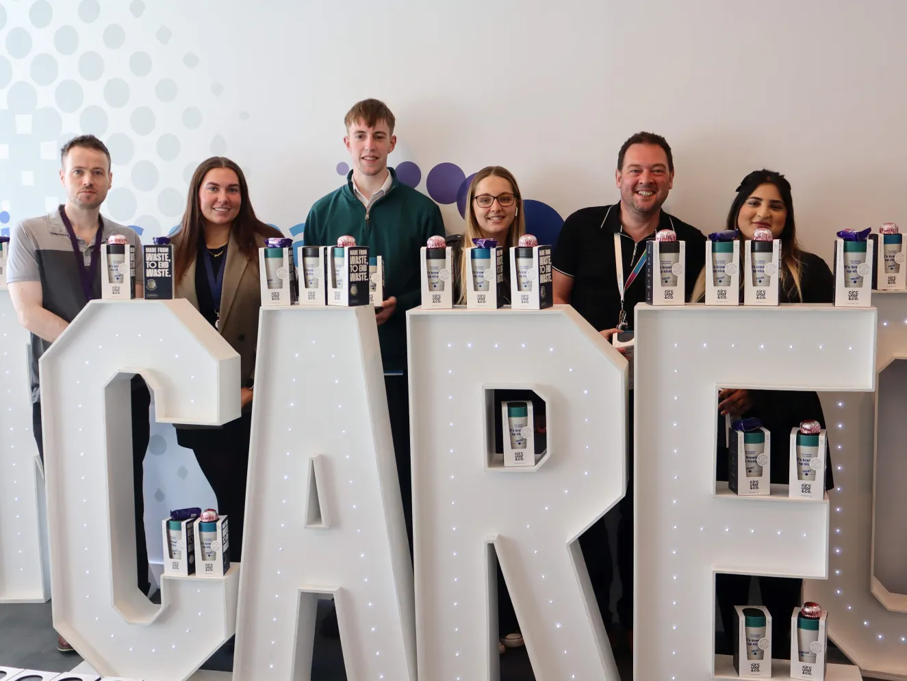 Six colleagues standing behind large floor-standing letters spelling out the world CARE