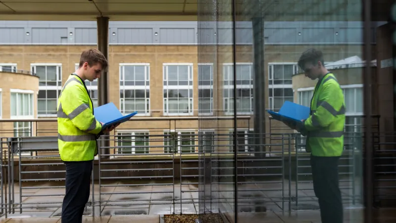Person in a high-visibility jacket reviewing documents beside a glass building, with their reflection visible in the window.