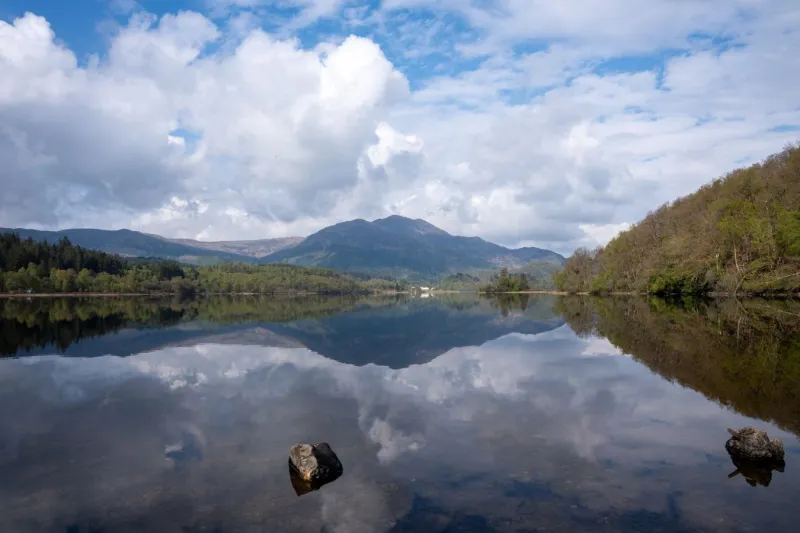 Loch Achray shot from low down. Still water in foreground with blue sky and clouds reflected, mountains behind