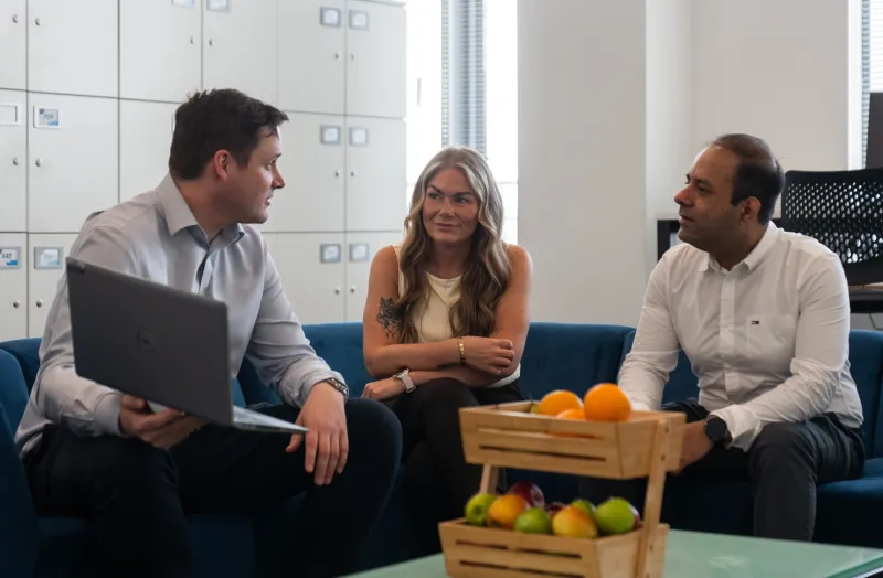 A woman and two men sitting talking in a breakout area with a basket of fruit in the foreground and lockers in the background