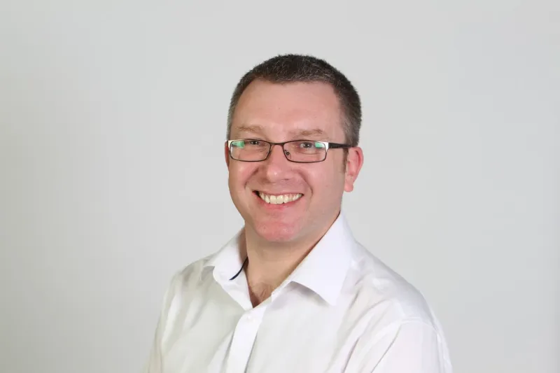 Headshot of Alex Lungley in a white shirt with no tie, smiling