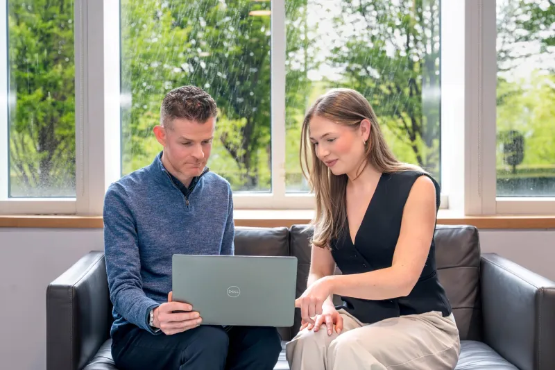 A man and woman looking at a laptop while seated side-by-side on a sofa in front of a window with trees outside.