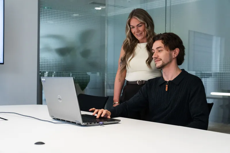 Woman standing behind man seated as both look at laptop screen with glass partition office behind them