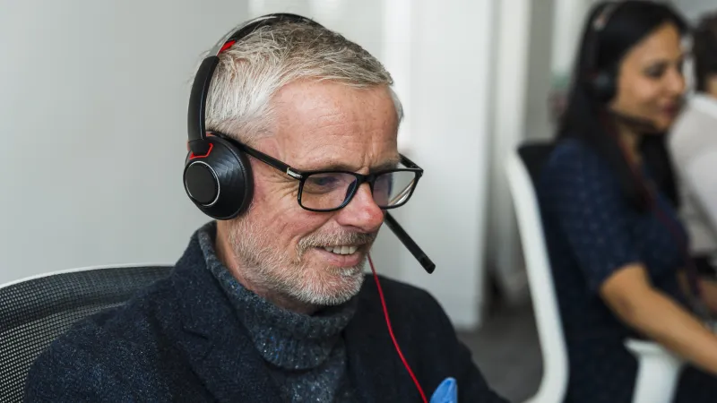 Man wearing headset smiling in customer service setting, with woman wearing headset in background