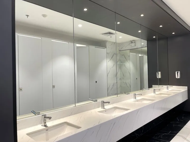 Row of modern white sinks in public toilet with cubicles reflected in mirror wall above