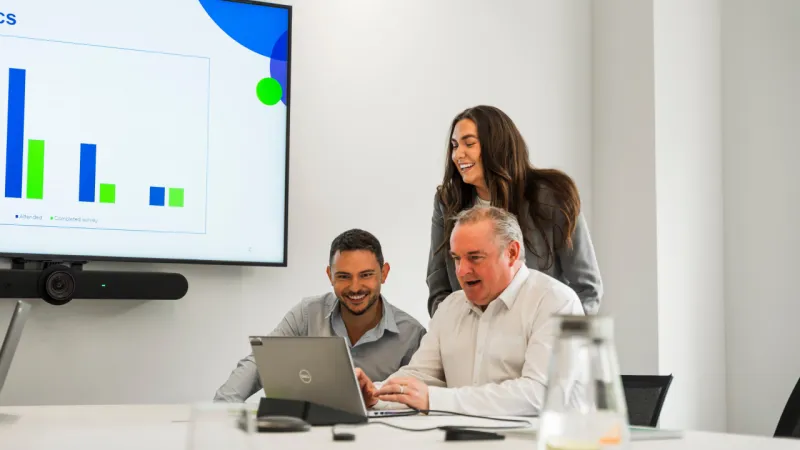 Three colleagues reviewing data on a laptop in a meeting room with a chart displayed on a screen