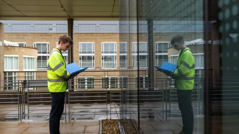 Person in a high-visibility jacket reviewing documents beside a glass building, with their reflection visible in the window.