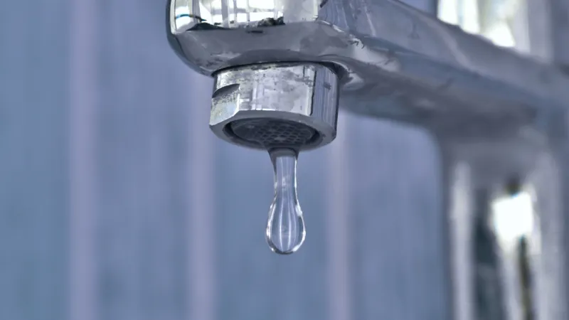 Close-up of a chrome tap with a single droplet of water hanging from the spout, set against a softly blurred blue background.