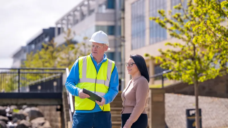 Two people talk outside near a waterfront development, with one wearing a hard hat and high-visibility vest while holding a tablet.