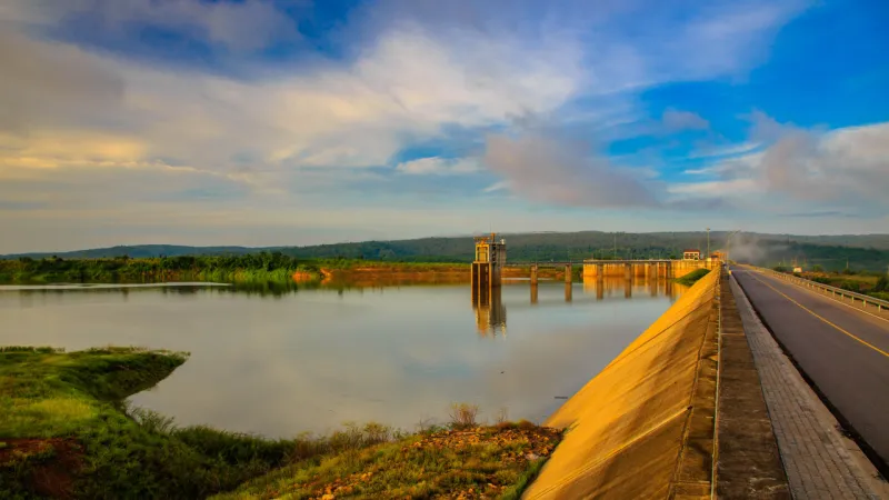 Dam and reservoir with calm water, a road along the dam wall, and rolling hills beneath a partly cloudy sky.