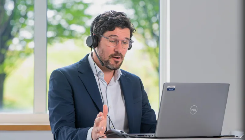 A man in a suit with no tie conducting a webinar at a laptop
