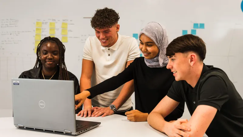Four young people of mixed ethnicity smiling and looking at a laptop in Business Stream office
